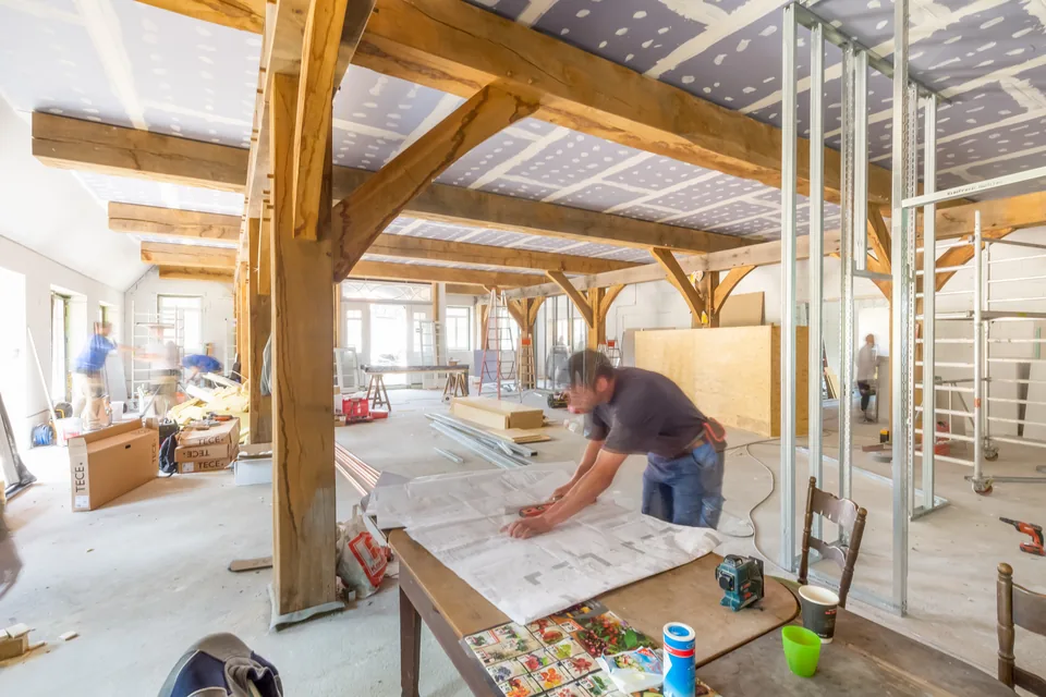 Baustelle im Dachgeschoss: Holzbalken, Trockenbau und ein Handwerker an einem Tisch mit Plänen, Gesicht unkenntlich gemacht