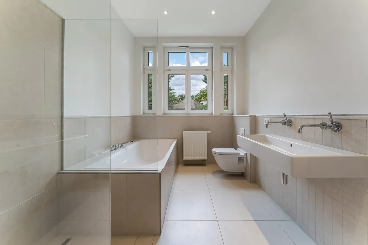 Nachher: Helles Badezimmer mit Badewanne, Glaswand und Doppelwaschbecken, Fensterblick mit blauem Himmel