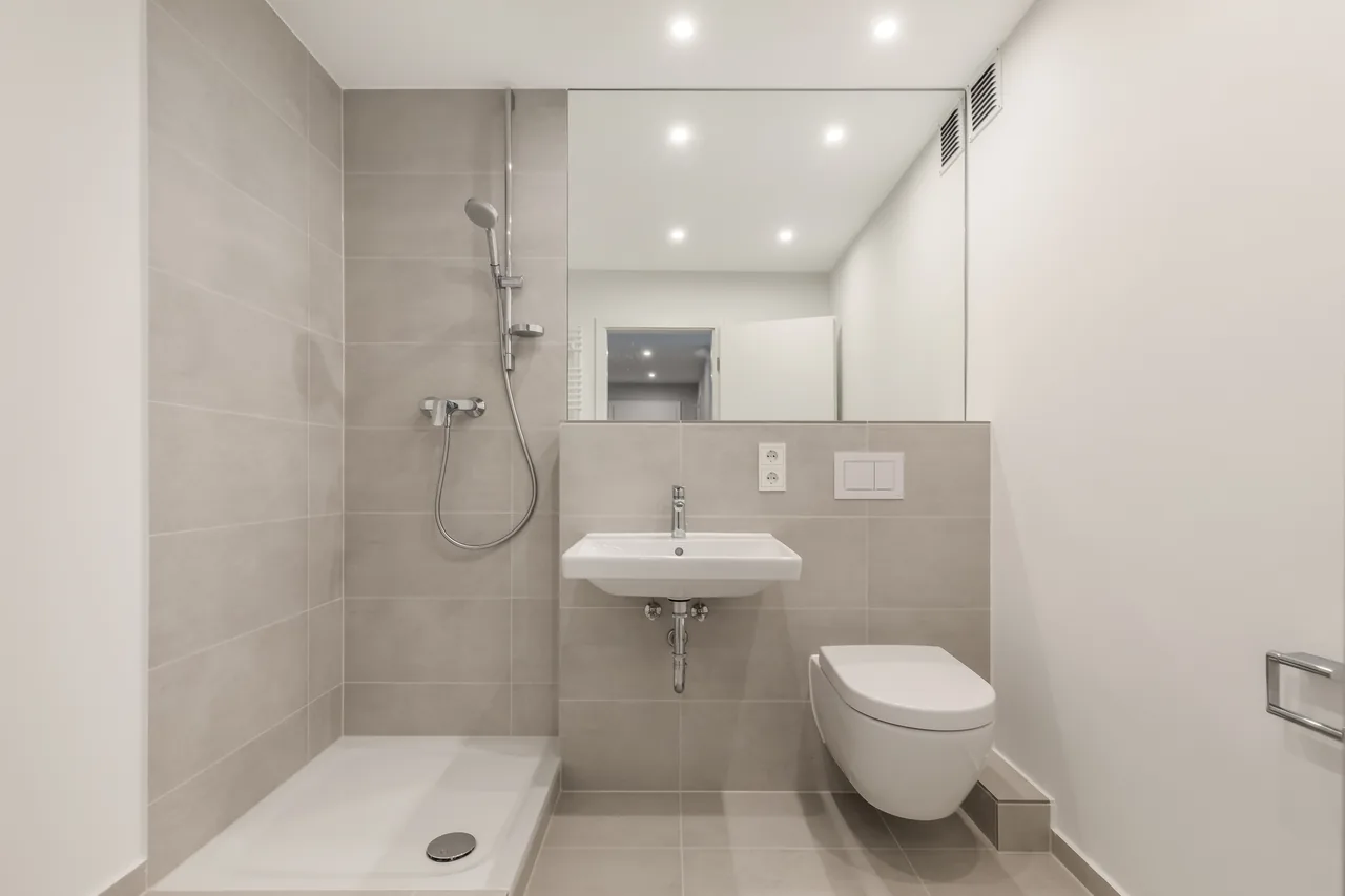 Nachher: dasselbe Bad mit grauen Fliesen, Dusche, Spiegel, Waschtisch und Wand-WC