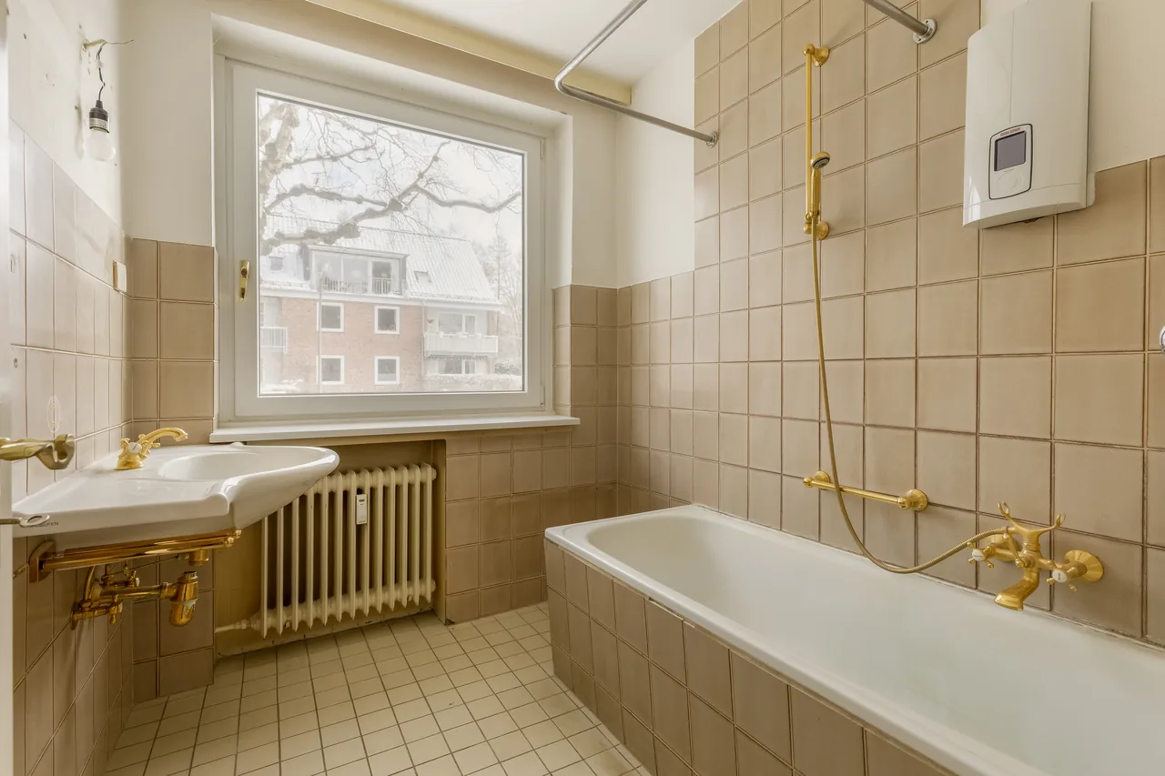 Vorher: Bad mit beigefarbenen Fliesen, Badewanne, goldfarbenen Armaturen und Heizkörper unter dem Fenster