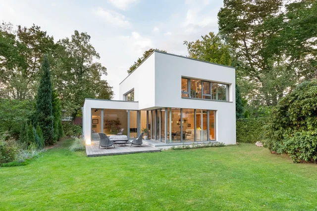 Moderne weiße Villa mit Flachdach und großen Glasfronten, Terrasse mit Sitzgruppe, Abendlicht im Inneren